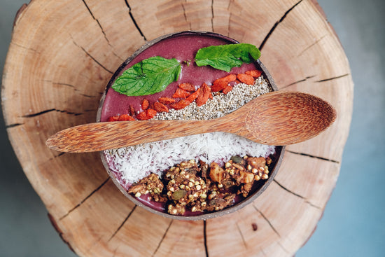 Smoothie bowl, smoothie rose orné de feuilles vertes, graines rose, copaux de noix de coco, et céréales. Une cuillère en bois est posée au dessus du bol.