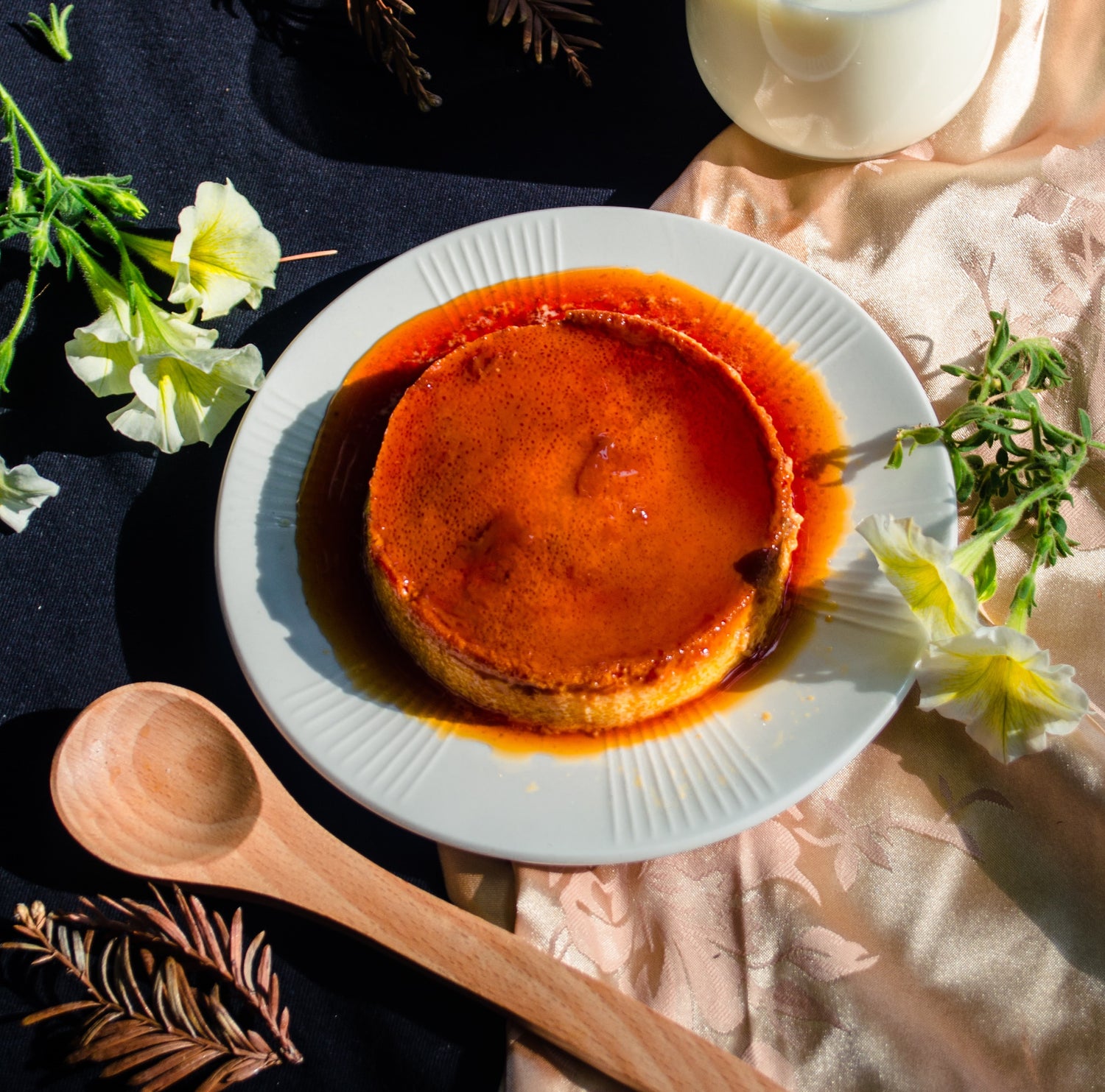Flan au caramel sur une assiette avec des fleurs à côté en décoration et une cuillère en bois