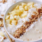 Smoothie bowl avec un smoothie jaune, des morceau de pomme, noix de coco, graines et céréales