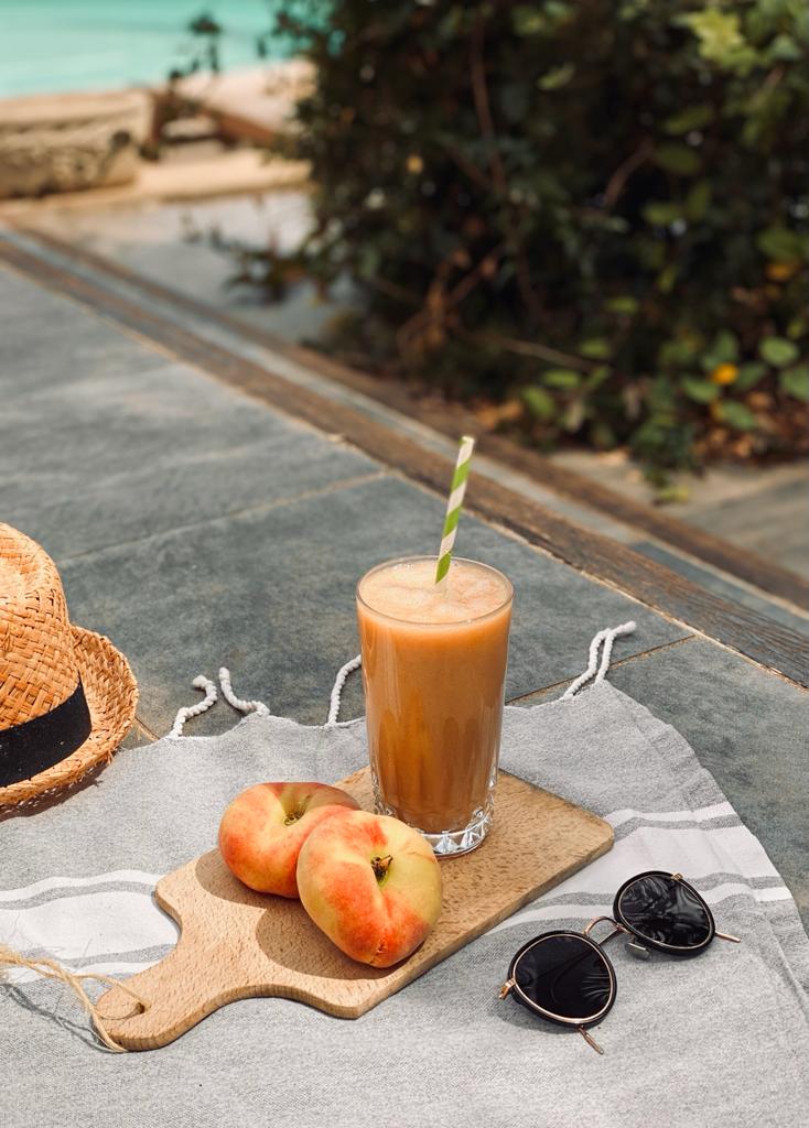 Verre à smoothie orange, accompagné de pêche sur une planche en bois sur une terrasse avec des lunettes de soleil. Ambiance vacances